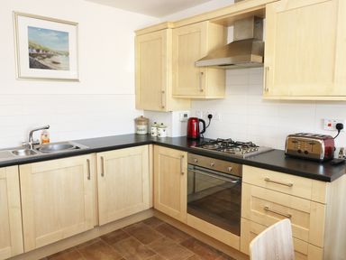 A kitchen with wooden cabinets black countertops a stainless steel sink gas stove oven toaster kettle and a framed painting at 5 Chandlers Yard in Burry Port