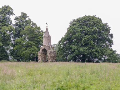 A grassy field with a small stone tower structure and large trees in the background at The Milking Barn in Yeovil Somerset