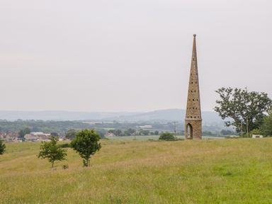 A grassy field with a tall conical stone monument and scattered trees near a village at The Milking Barn in Yeovil Somerset
