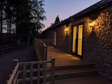 An outdoor wooden deck with railing and stairs attached to a stone building with lit wall lamps at The Milking Barn in Yeovil Somerset