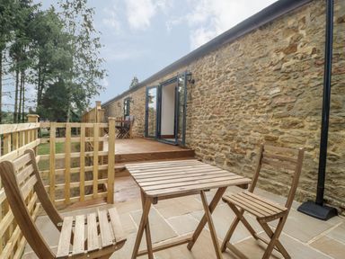 An outdoor patio area with a wooden table and two chairs next to a stone wall and wooden deck at The Milking Barn in Yeovil Somerset