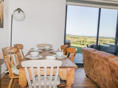A dining area with a wooden table and chairs set with bowls next to a brown sofa and a large window showing an outdoor patio at Heron Lodge Castle Farm in Wedmore