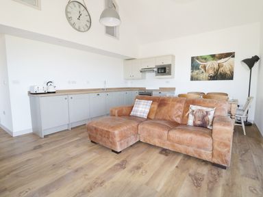 An open plan kitchen and living area with a brown leather sofa wooden dining table and chairs wall clock and kitchen appliances at Heron Lodge Castle Farm in Wedmore