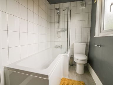 A bathroom with a bathtub shower combination white tiles a toilet a window and a yellow bath mat at 2 Hillside Cottages in Felixstowe Suffolk