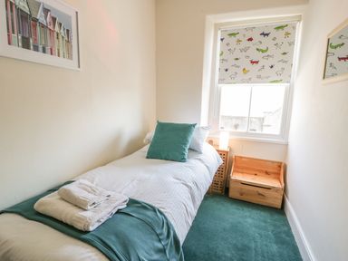 A small bedroom with a single bed with white bedding and teal pillow and throw a wooden chest and bedside table by a window with animal patterned blind at Chwech in Menai Bridge