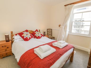 A bedroom with a double bed red cushions and towels on the bed a wooden nightstand with a lamp a chair and a window with curtains at Chwech in Menai Bridge