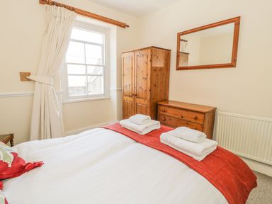 A bedroom with a bed made with white sheets and red runner with towels on top beside a wooden wardrobe and chest of drawers at Chwech in Menai Bridge