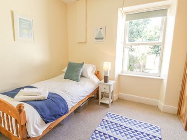 A bedroom with a single wooden bed white bedding blue blanket and pillow bedside table with lamp window and patterned rug at Chwech in Menai Bridge