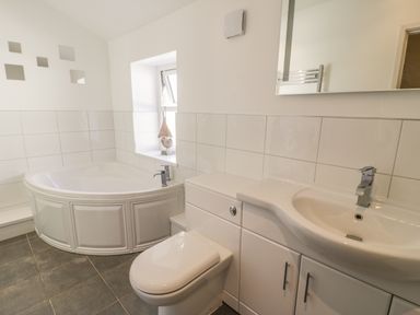 A bathroom with a corner bathtub toilet sink white cabinets and a small window at Chwech in Menai Bridge