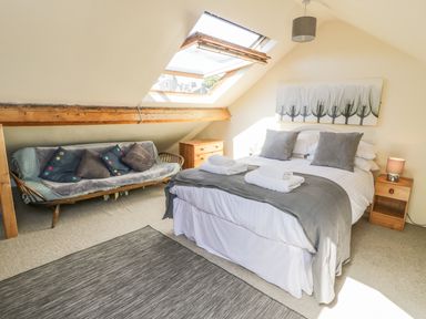 A bedroom with a double bed, grey pillows, folded towels, a wooden side table with a lamp, a wooden chest of drawers, a sofa with cushions under a slanted ceiling window at Chwech in Menai Bridge