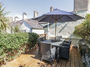 An outdoor wooden deck with a table six chairs and a large umbrella surrounded by plants at Chwech in Menai Bridge