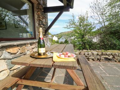 A wooden picnic table with a bottle of champagne two glasses and a plate of macarons outside a stone house garden and hills visible