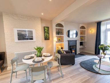 A dining table with four chairs and flowers beside a sitting area with an armchair fireplace and shelves at Harbour View Cottage in Watchet