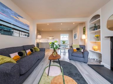 An open plan living room with gray sofas, a coffee table with flowers, a dining area, and a kitchen at Harbour View Cottage in Watchet