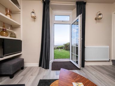 A room with an open glass door leading to a garden with grass and a birdbath at Harbour View Cottage in Watchet