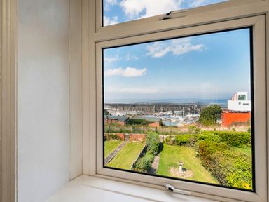 A view through a window showing a garden with bushes and a birdbath and a marina with boats beyond at Harbour View Cottage in Watchet