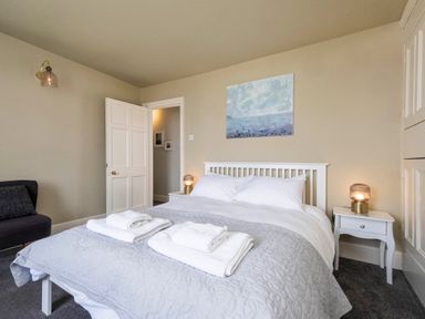 A bedroom with a bed with folded towels bedside tables with lamps a chair and a painting on the wall at Harbour View Cottage in Watchet