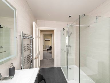 A bathroom with a glass shower enclosure a towel radiator a sink and a doorway leading to a carpeted room at Harbour View Cottage in Watchet