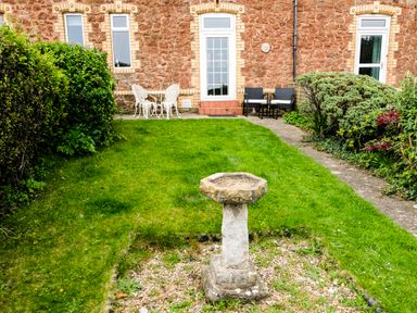 A garden with a stone birdbath green grass bushes and outdoor chairs by a brick house at Harbour View Cottage in Watchet