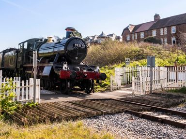 A steam locomotive stopped at a railway crossing with white fences and houses in the background at Harbour View Cottage in Watchet