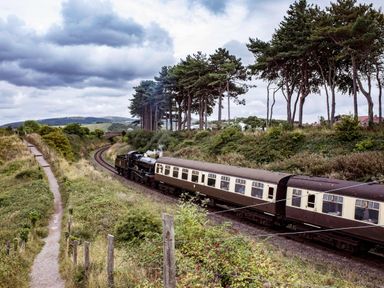 A steam train traveling on a curved railway track next to a dirt path and trees at Harbour View Cottage in Watchet