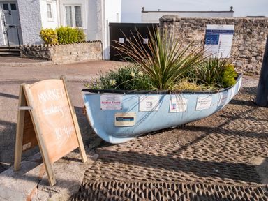 A small boat filled with plants on a cobblestone street next to a wooden sign that says market today 10 to 4 pm at Harbour View Cottage in Watchet