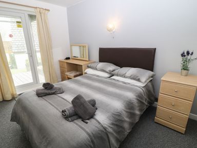 A bedroom with a double bed gray bedding towels on bed wooden dresser bedside table with flowers and window with curtains at Running Waters in Callington