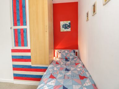 A small bedroom with a single bed with patterned bedding a wooden wardrobe and a red accent wall at Swanage Bay Apartment in Swanage