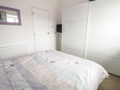 A bedroom with a bed covered by a patterned bedspread a white door a wall-mounted shelf with a TV and a large white wardrobe at Swanage Bay Apartment in Swanage