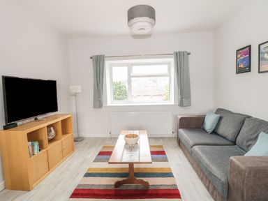 A living room with a sofa a wooden coffee table a striped rug a wooden cabinet with a TV and a window at Swanage Bay Apartment in Swanage