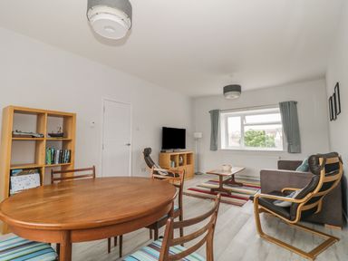 A living room with a wooden dining table and chairs a bookshelf a sofa an armchair a coffee table a TV and a window with curtains at Swanage Bay Apartment in Swanage
