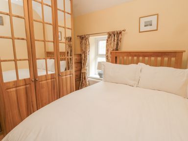 A bedroom with a wooden bed white bedding wooden wardrobes and a window with floral curtains at Red Squirrel Cottage 5 Biddlestone in Rothbury