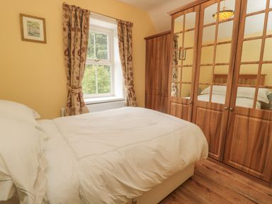 A bedroom with a bed draped in white linens next to a window with curtains and a wooden wardrobe with mirrored doors at Red Squirrel Cottage 5 Biddlestone in Rothbury