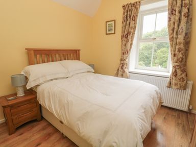 A bedroom with a wooden bed a bedside table with a lamp and a window with curtains at Red Squirrel Cottage 5 Biddlestone in Rothbury