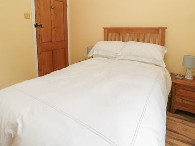 A bedroom with a wooden bed with white bedding a wooden door and a wooden bedside table with a lamp and remote at Red Squirrel Cottage 5 Biddlestone in Rothbury