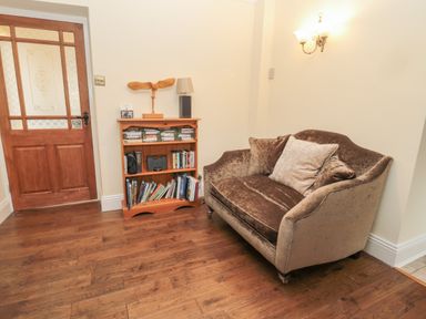 A small sitting area with a brown cushioned loveseat next to a wooden bookshelf and a wooden door at Red Squirrel Cottage 5 Biddlestone in Rothbury