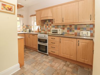 A kitchen with wooden cabinets a tiled backsplash a stove a microwave a toaster and two windows at Red Squirrel Cottage 5 Biddlestone Rothbury