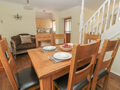 A dining area with a wooden table set with plates and cutlery six wooden chairs a brown sofa an open archway leading to a kitchen and a staircase with white spindles at Red Squirrel Cottage 5 Biddlestone in Rothbury