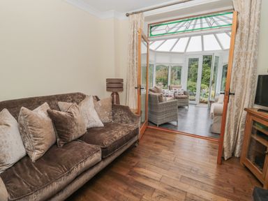 A living room with a brown sofa and pillows open to a conservatory with wicker chairs and a garden view at Red Squirrel Cottage 5 Biddlestone in Rothbury