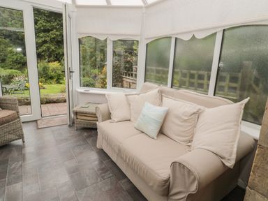 A conservatory with a beige sofa and cushions a wicker chair a small wicker table and glass doors leading to a garden at Red Squirrel Cottage 5 Biddlestone Rothbury