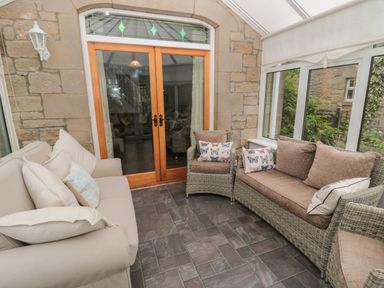 A conservatory with a beige sofa and wicker chairs with cushions by wooden double doors at Red Squirrel Cottage 5 Biddlestone Rothbury