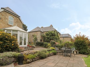 An outdoor patio area with a metal table and chairs surrounded by plants and stone buildings at Red Squirrel Cottage 5 Biddlestone in Rothbury