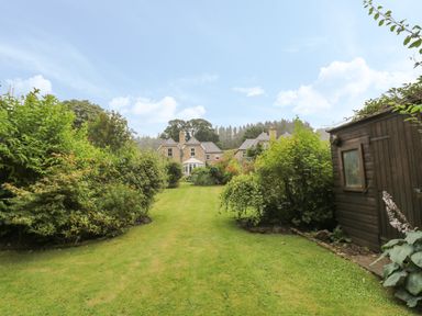 A garden with a lawn running between bushes and trees with a wooden shed on the right and two houses in the background at Red Squirrel Cottage 5 Biddlestone in Rothbury