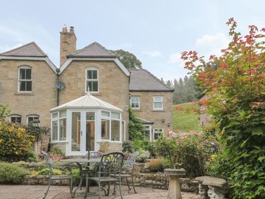 An outdoor patio with a round metal table and chairs surrounded by plants and flowers at Red Squirrel Cottage 5 Biddlestone in Rothbury