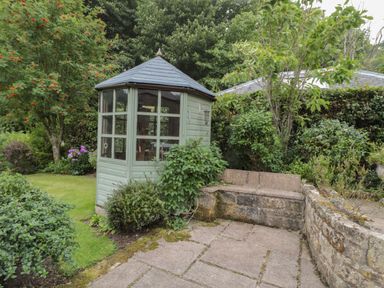 A garden with a small green wooden gazebo and stone seating area surrounded by bushes and trees at Red Squirrel Cottage 5 Biddlestone Rothbury