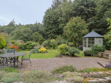 A garden with a metal table and chairs on a paved area and a small gazebo surrounded by plants and trees at Red Squirrel Cottage 5 Biddlestone in Rothbury