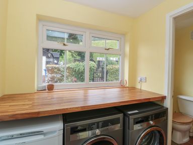 A laundry room with a wooden countertop above a washing machine and dryer next to a window and a doorway leading to a bathroom at Red Squirrel Cottage 5 Biddlestone in Rothbury