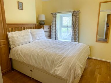A bedroom with a double bed, bedside table with lamp, window with curtains, wall mirror and wardrobe at Red Squirrel Cottage 5 Biddlestone Rothbury