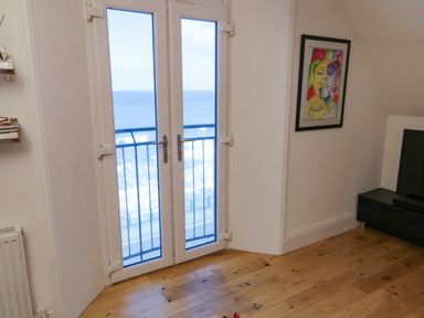 A room with wooden floor a glass door opening to a balcony overlooking the sea and a wall with a colorful framed painting at Flat 3 Peacehaven in Sandsend near Whitby