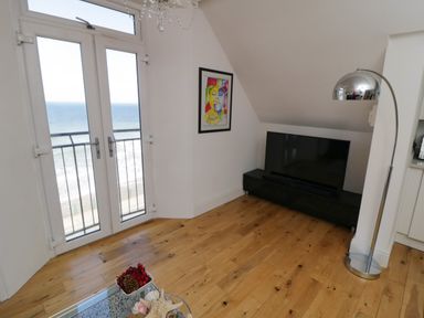 A room with wooden floor a large TV on a stand a chrome floor lamp colorful wall art and double glass doors with sea view at Flat 3 Peacehaven Sandsend near Whitby